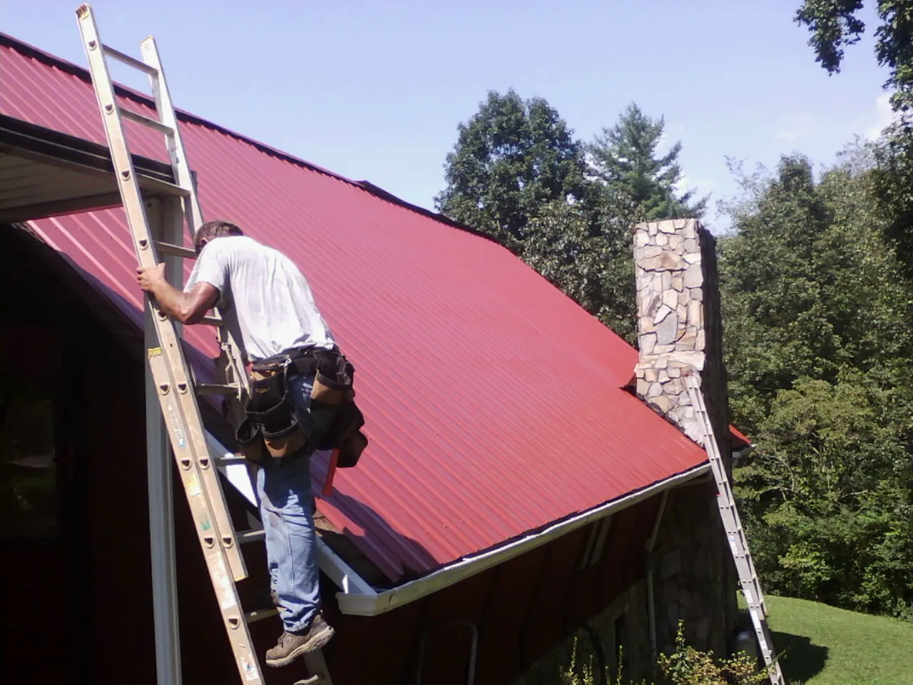 Roofer on a ladder beside a red metal roof and stone chimney — Soffit & Fascia Repair by Roofing Brothers in Eden